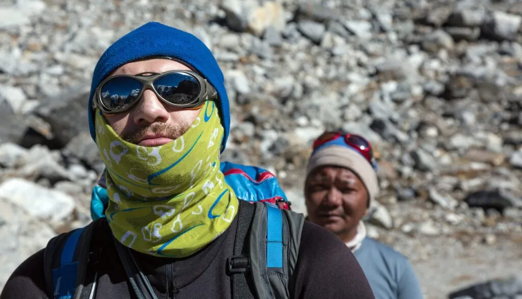 trekker w czapce, okularach przeciwsłonecznych i chuście na twarzy, obok przewodnik na kamienistym szlaku